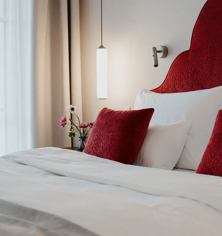 Stilvoller Schlafbereich mit rotem Samtkopfteil, weißen Bettlaken, roten Zierkissen und Blumen im Grand Elisabeth Hotel in Bad Ischl.