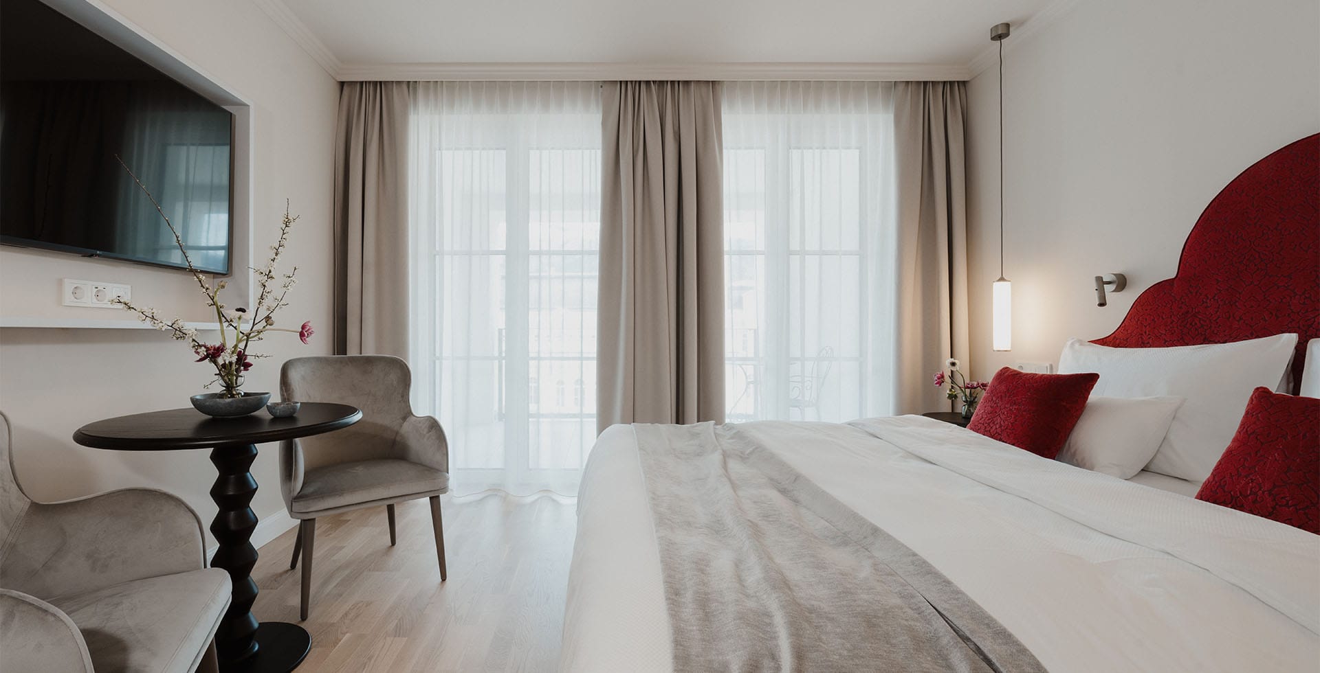 Gemütliches Combi Classic Doppelzimmer mit rotem Kopfteil, edlen Kissen und Sitzbereich am Balkon im Grand Elisabeth Hotel in Bad Ischl.