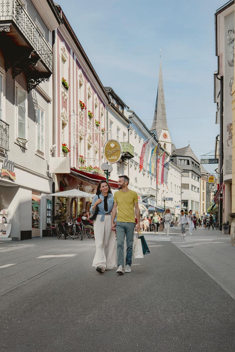 Ein Paar geht durch die Innenstadt von Bad Ischl mit Einkaufstüten, umgeben von Sehenswürdigkeiten und Geschäften