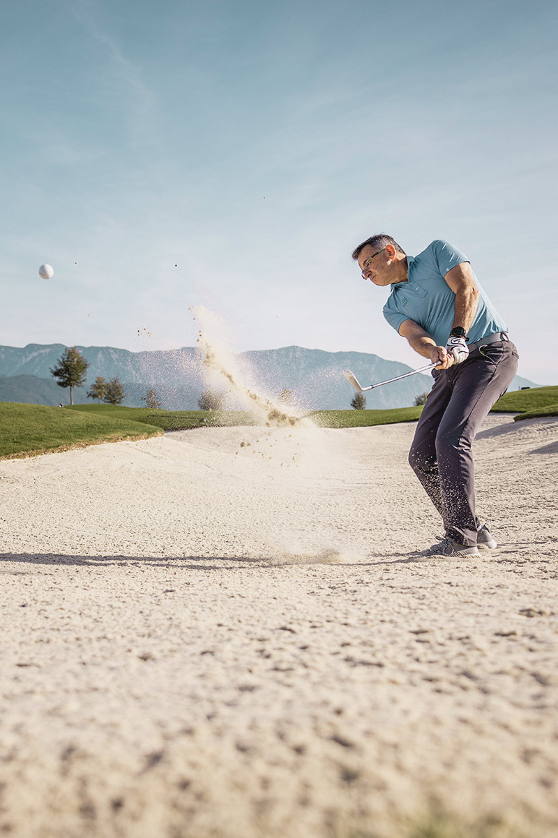 In einer Sandgrube auf dem Golfplatz schlägt ein Mann den Ball, im Hintergrund das Bergpanorama des Salzkammerguts