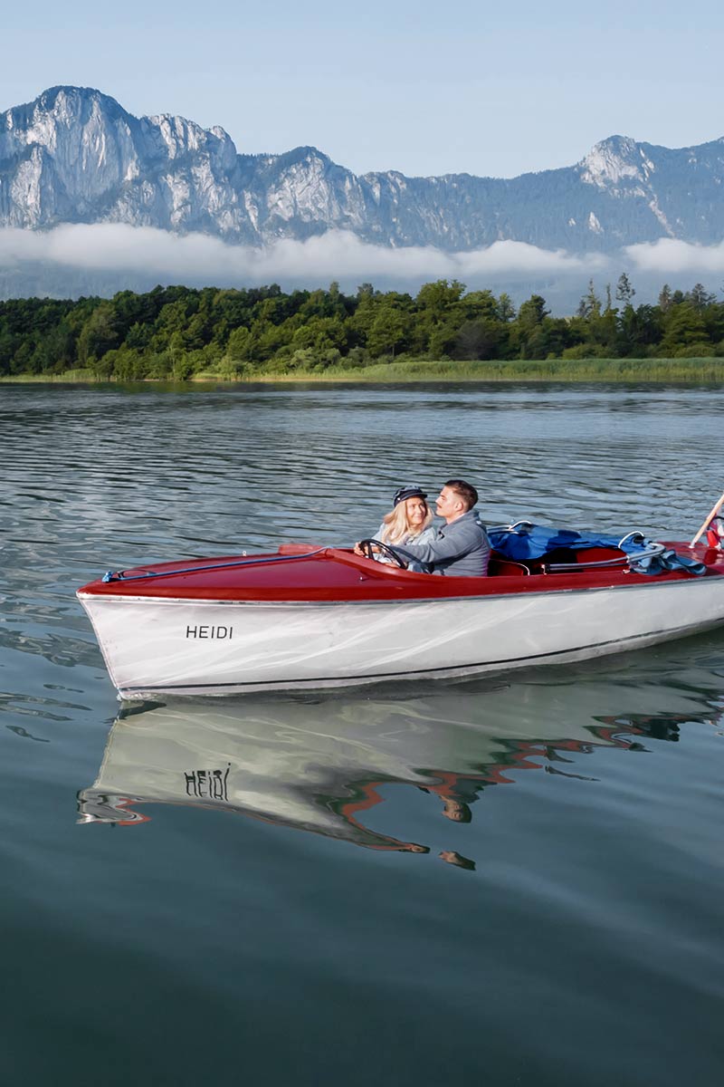 Ein Paar fährt bei sonnigem Wetter mit einem Motorboot über den Wolfgangsee, im Hintergrund sind Berge