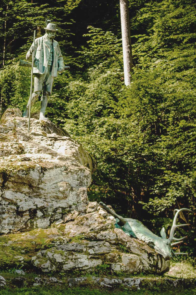 Am Waldrand im Salzkammergut steht eine Jägerfigur auf einem Stein, zu ihren Füßen eine Hirschfigur