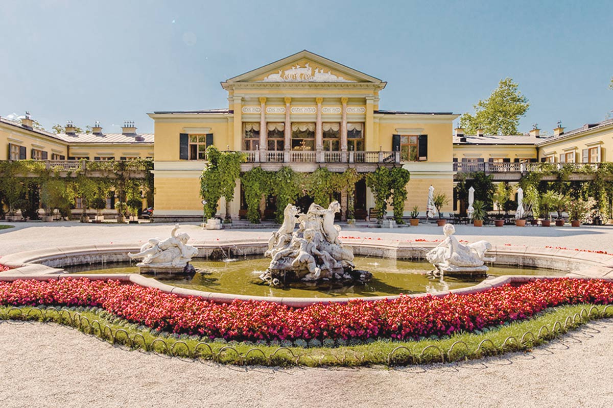 Aufnahme der Kaiservilla in Bad Ischl, davor ein Brunnen mit kunstvollen Statuen, umgeben von Blumenbeeten